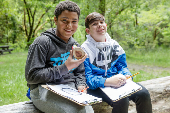 Two smiling boys sit on a log holding clipboards. One holds a cross-section of a tree.