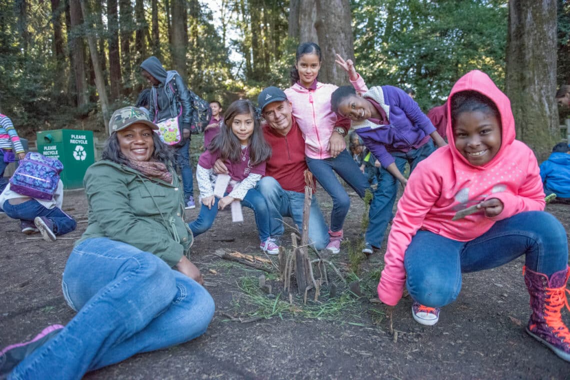 A group of smiling children and adults sit and kneel on the floor of a redwood forest