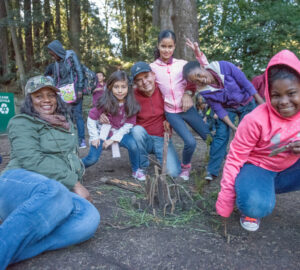 A group of smiling children and adults sit and kneel on the floor of a redwood forest