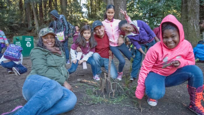 A diverse group of smiling children and adults of color sit and kneel on the floor of a redwood forest. Oakland students explored the redwoods at Roberts Regional Park
