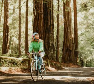 A woman wearing a bright green shirt and a bike helmet rides a bike down a paved path in the redwoods