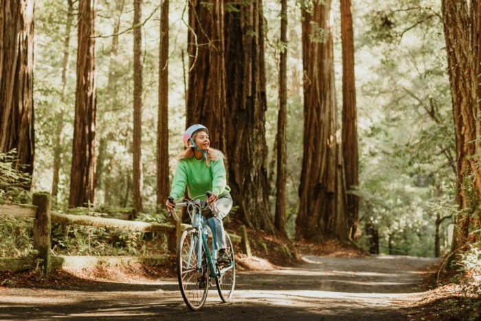 A woman wearing a bright green shirt and a bike helmet rides a bike down a paved path in the redwoods