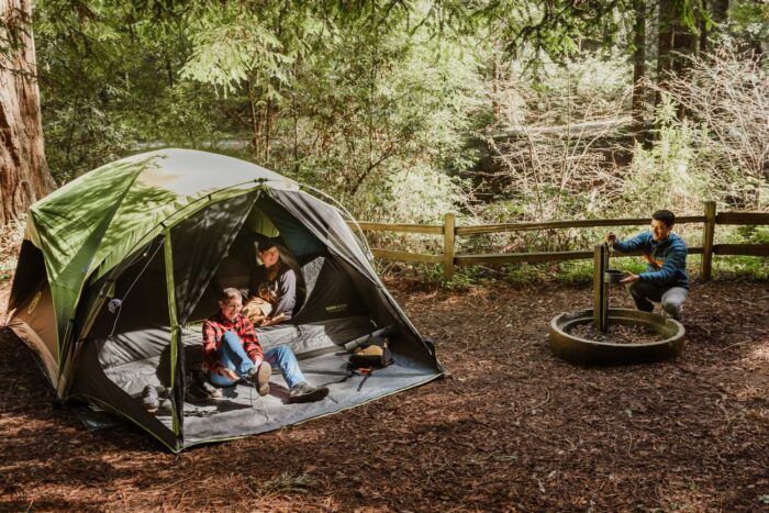 At a shady campsite, two people sit inside a green tent while a man kneels at a water spigot to fill a cooking pot