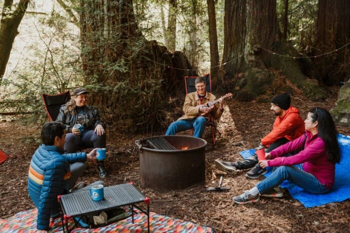 A person strums a guitar at a campfire ring. Four people sit around the fire with mugs beneath large sunlit redwood trees.