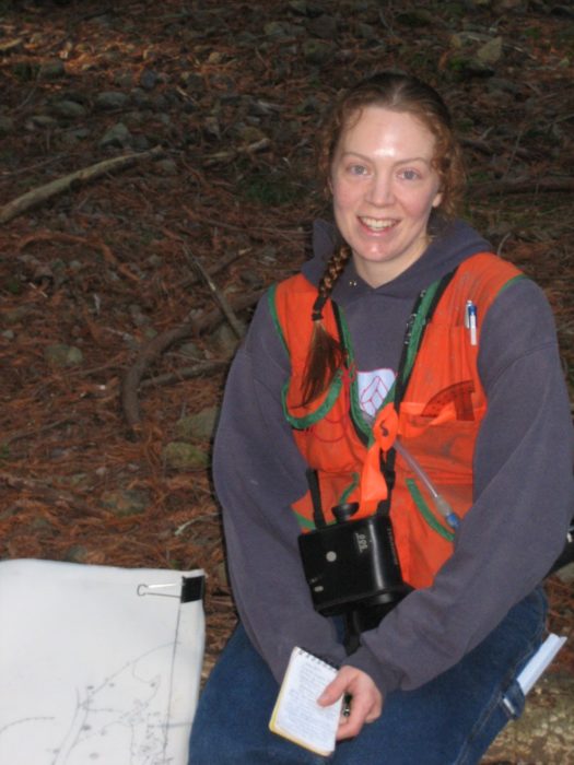 A red-haired women wearing a grey hoodie and an orange vest sits on a bed of fallen leaves