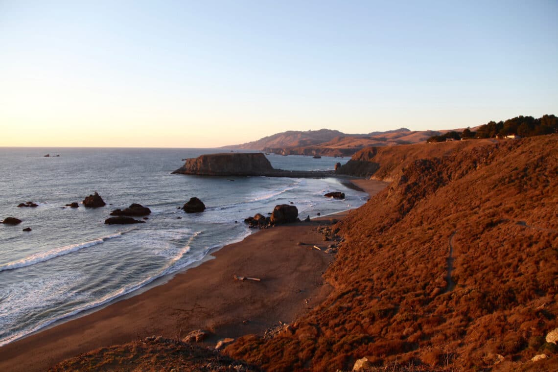 Bluffs lead down to a dark sand beach at sunset. Rock formations rise from the ocean.