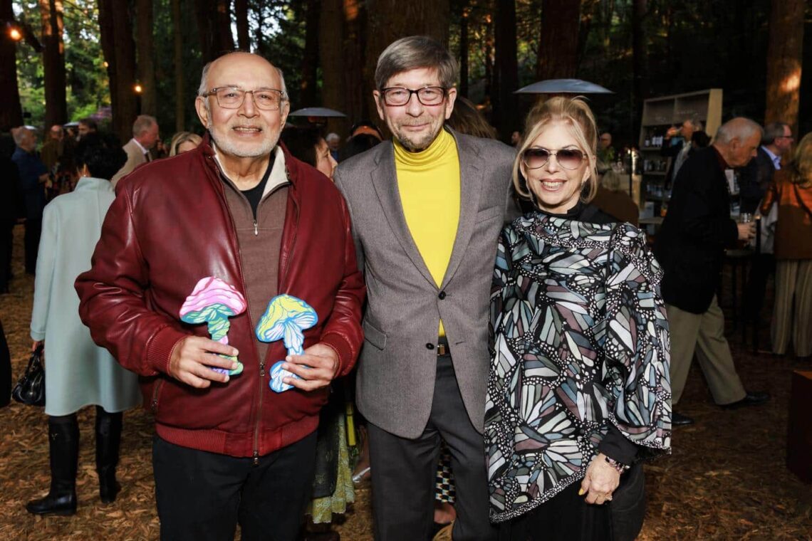 Two men and a woman who are guests at the event. The older man on the left, wearing glasses, has on a red leather bomber jacket, while the man in the middle has on a yellow turtleneck underneath his grey blazer - a nod to the banana slug. A blonde woman wearing sunglasses with yellow painted nails stands on the right.