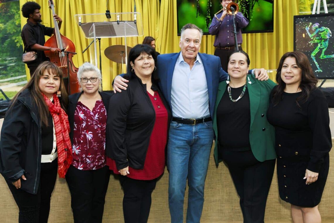Five women and one man stand together to pose for a photo in front of the stage with musicians standing behind them.