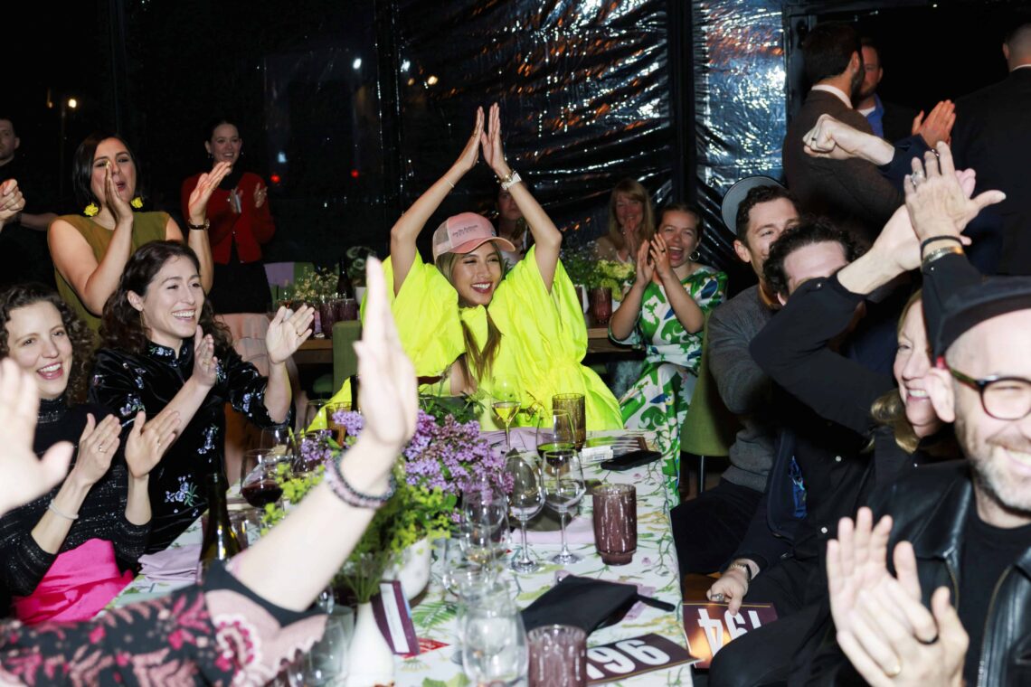 A woman in a bright banana slug yellow jacket cheers and claps with her hands above her head at the end of a table surrounded by clapping guests