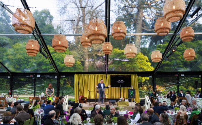a festive crowd seated at tables in an open-air tent beneath the trees. A man addresses them from the stage.