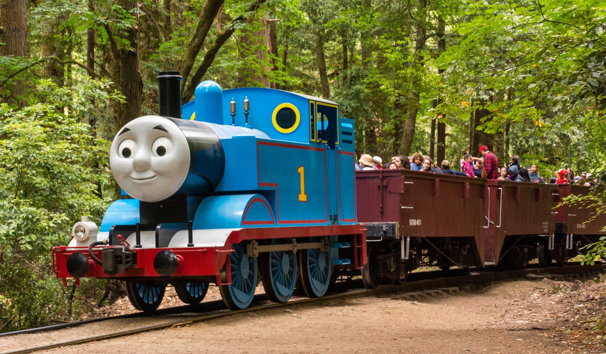 A bright blue locomotive that looks like Thomas the Train Engine pulls an open-air passenger car filled with people