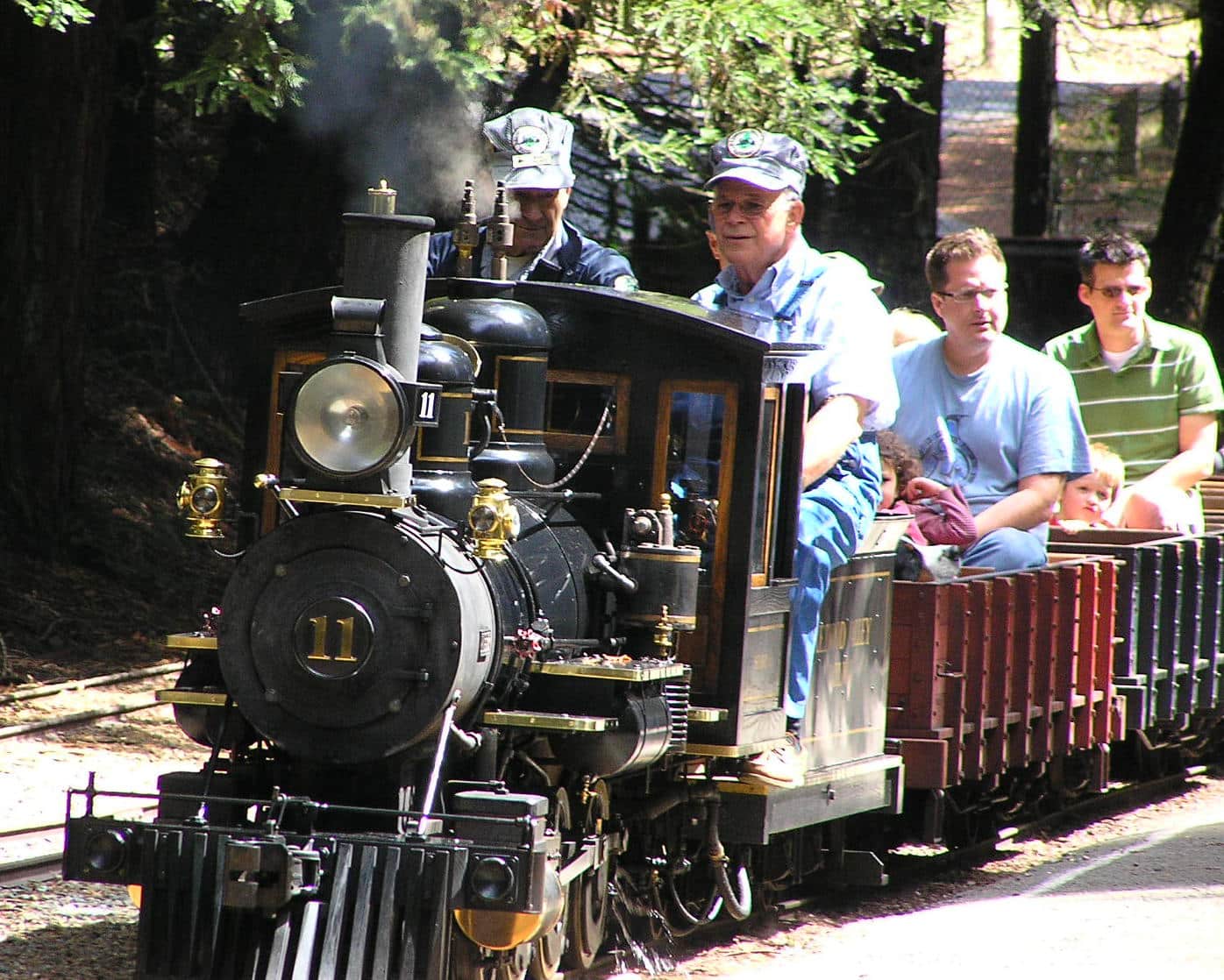 Two train engineers with striped caps drive a miniature steam train pulling open-air cars with parents and kids
