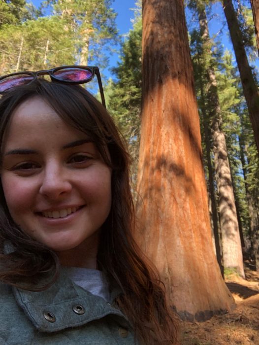 A woman smiling with sunglasses on her head and a giant sequoia tree in the background