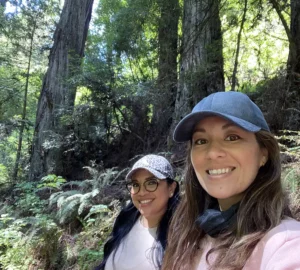 Two people smile in the foreground; a sunny redwood forest is in the background.