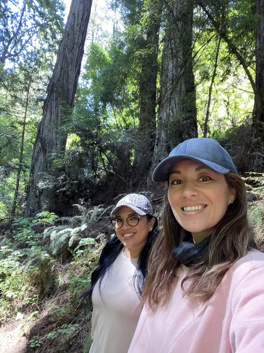 Two people smile in the foreground; a sunny redwood forest is in the background.