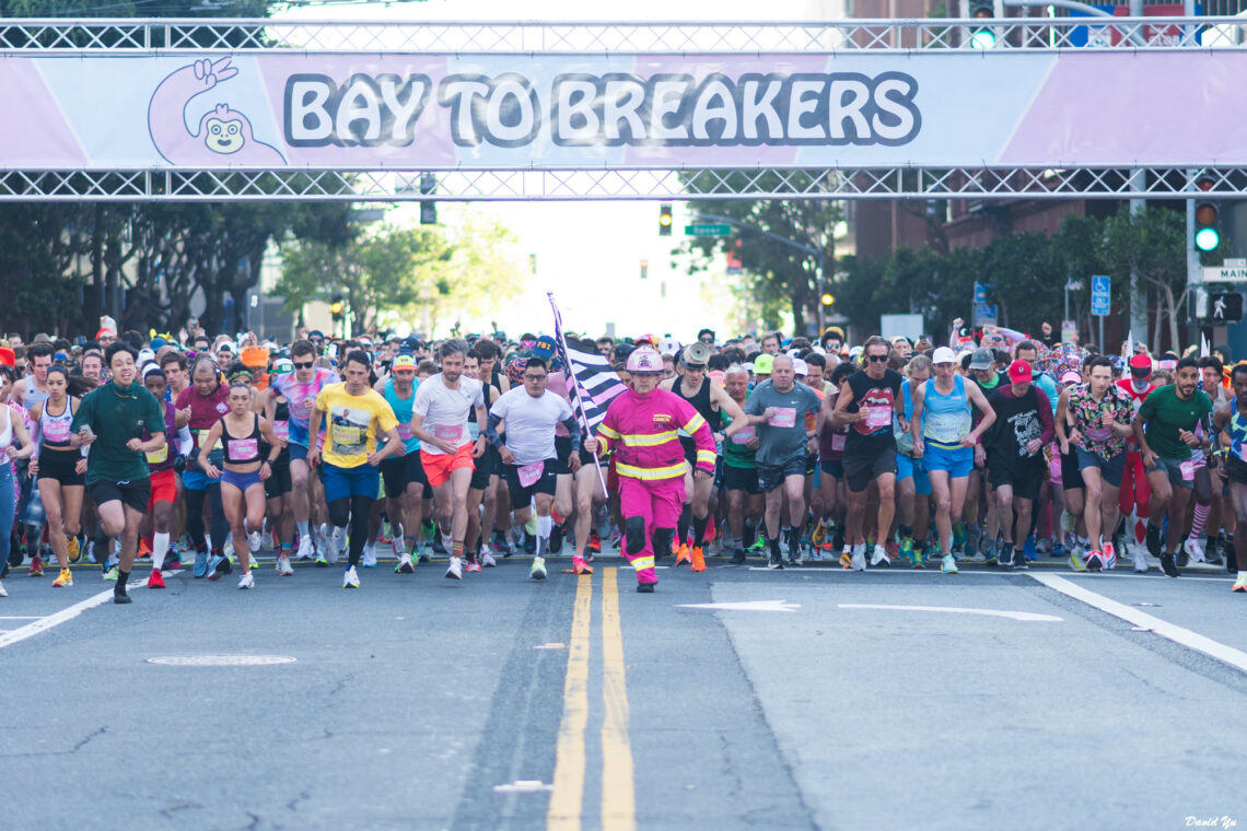 A large crowd of runners in colorful outfits takes off from the starting line of the Bay to Breakers race in San Francisco, under a banner that reads 'Bay to Breakers' with a cartoon hand-waving character.
