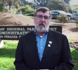 A man with a beard and glasses standing in front of a park sign with a car in the background