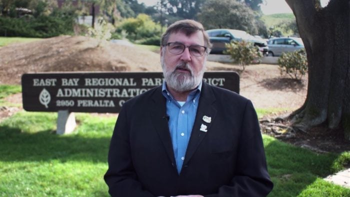 A man with a beard and glasses standing in front of a park sign with a car in the background