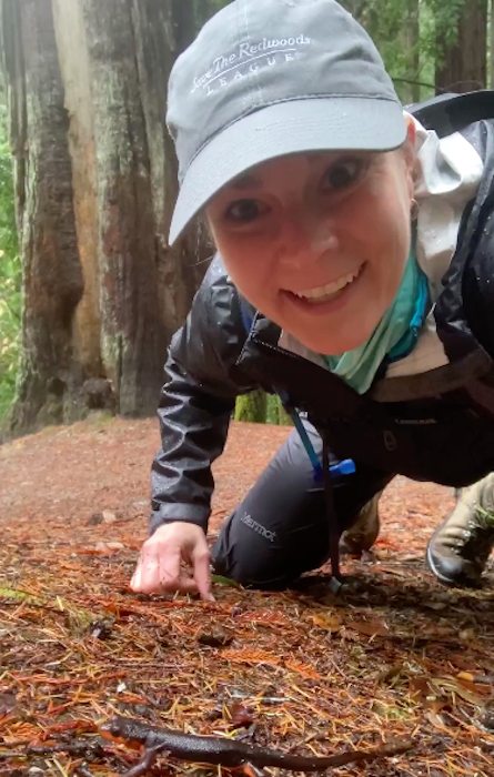 A woman wearing a baseball cap, crouching down on the forest floor with a newt