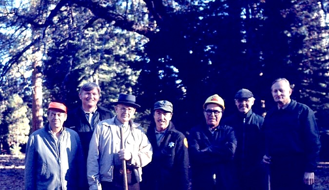 A group of men poses for the camera in a forest, some wearing helmets and park uniforms, others carrying rakes
