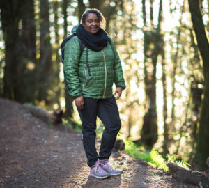 Square cropped photo of author Teresa Baker, a black woman, smiling outside on a wide trail. She wears a scarf, green puffy coat, dark pants and grey sneakers with pink accents.