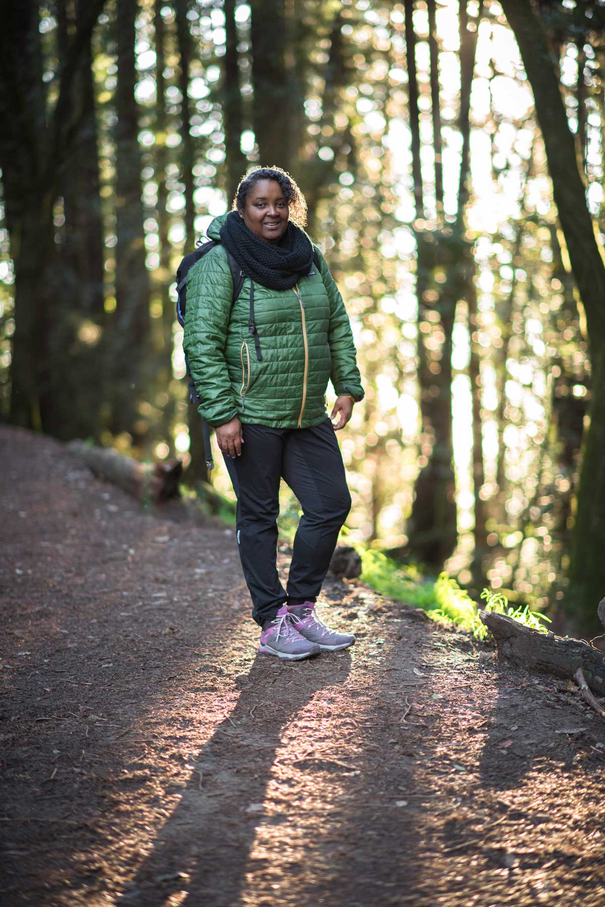Portrait photo of author Teresa Baker, a black woman, smiling outside on a wide trail. She wears a dark scarf, green puffy jacket, dark denims, and grey sneakers with pink accents. 