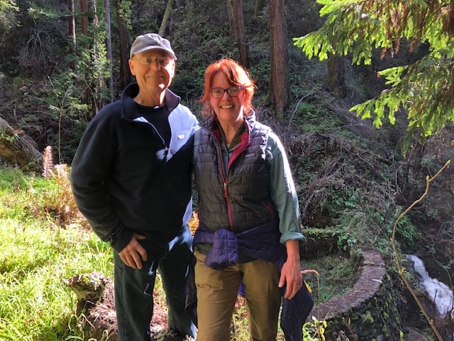 A man and a woman standing close together in the sunlight, with a forest in the background