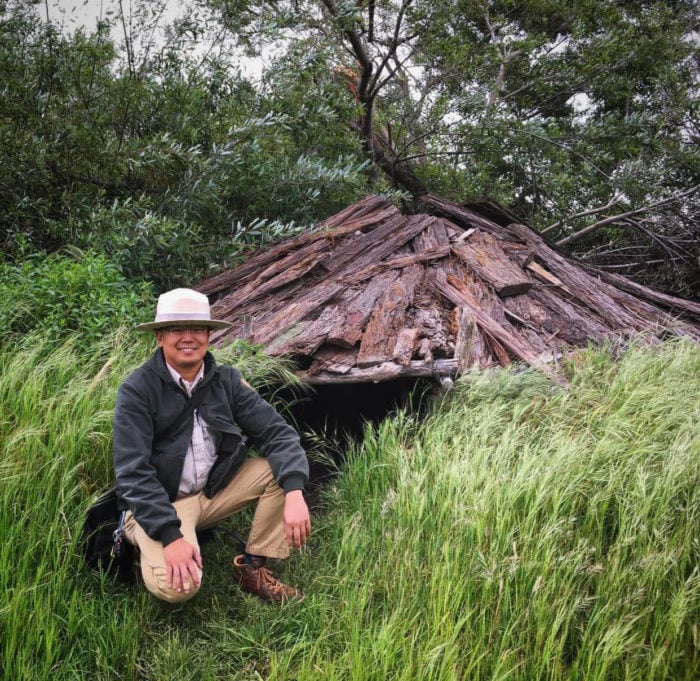 A park ranger kneels on green grass in front of a sweathouse with a redwood bark roof