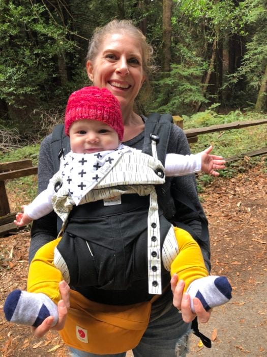 A woman smiling with a baby wearing a red hat in a sling, with redwood trees in the background