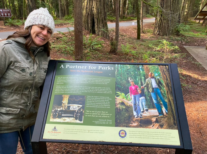 A woman wearing a beanie standing next to a park interpretive panel in a redwood forest