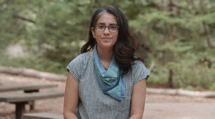 A woman wearing eyeglasses and a bandana around her neck, with greenery in the background