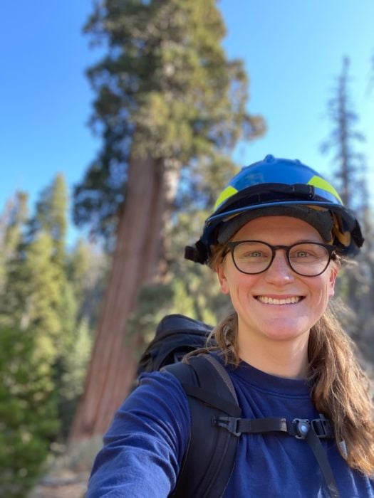 A woman wearing a hard hat and eyeglasses smiles in front of a giant sequoia tree.