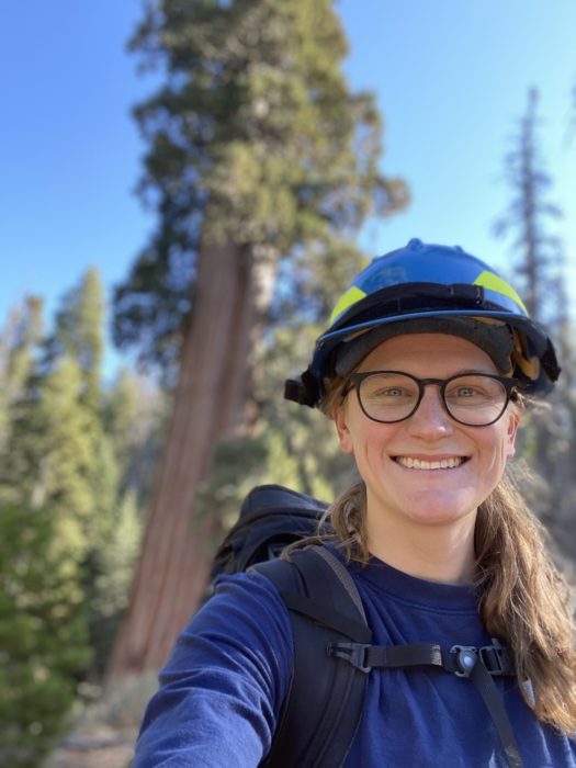 A woman wearing a hard hat and eyeglasses smiles in front of a giant sequoia tree.