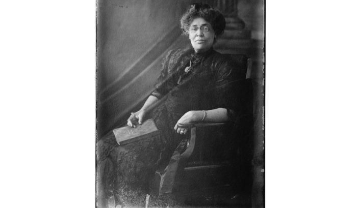 Oldl black-and-white photo of a woman sitting in a chair and holding a book.