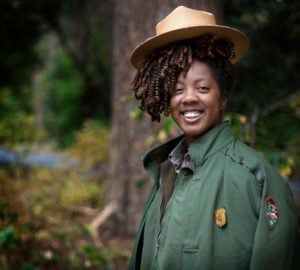 A smiling Black woman ranger in uniform with a forest in the background