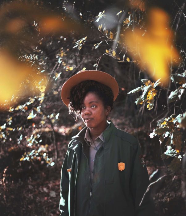 Image of a Black woman National Park Service ranger in uniform framed by yellow foliage in the foreground and foliage in the background