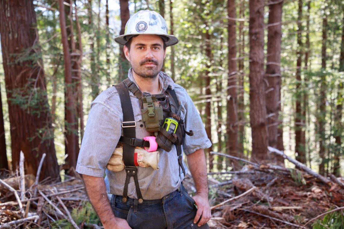 A man wearing a silver hard hat and a chest harness that holds various tools looks at the camera. Behind him is a redwood forest and a pile of small branches.