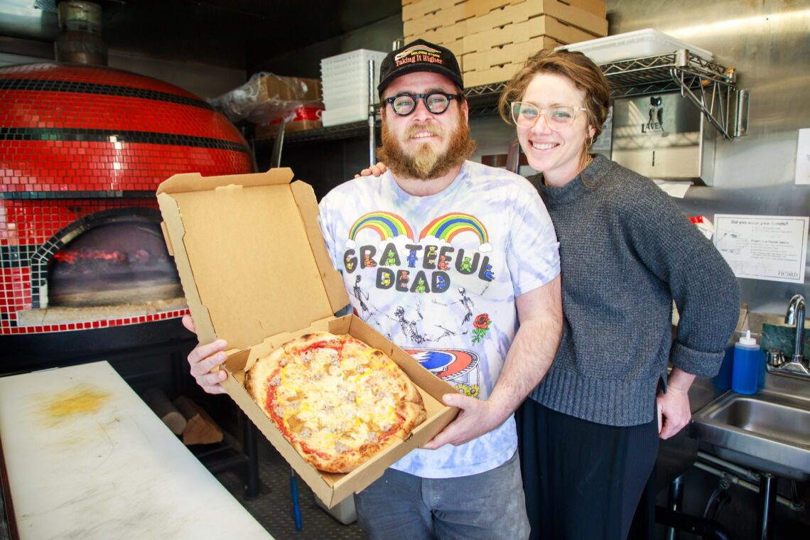 A man wearing a Grateful Dead t-shirt proudly holds an open pizza box with a pizza inside. A smiling woman stands next to him, in a small kitchen with a wood fired pizza oven