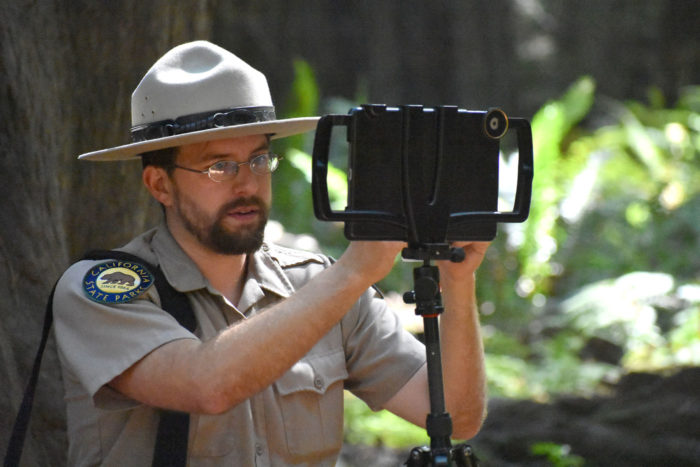 A bearded California State Parks naturalist interpreter wearing glasses and park ranger fatigues adjusts a tablet on a tripod in preparation for broadcasting a livestream to Facebook.