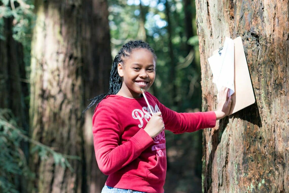 A student standing next to a tree smiles as she prepares to take notes