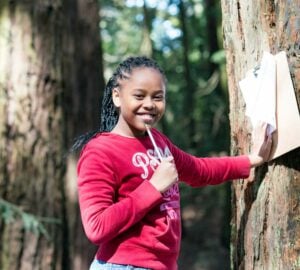 A student standing next to a tree smiles as she prepares to take notes