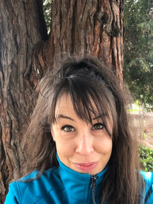 A brunette woman wearing a teal jacket and standing in front of a redwood tree