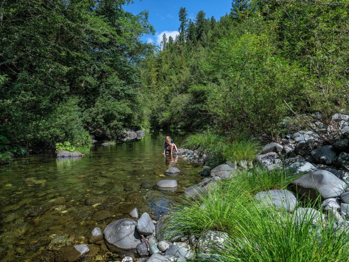 A woman sits on a rock along a river with clear water, surrounded by lush foliage and a blue sky