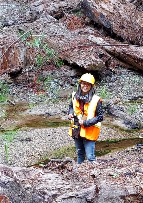 A woman wearing a yellow hard hat and orange and yellow vest, standing in a creek.
